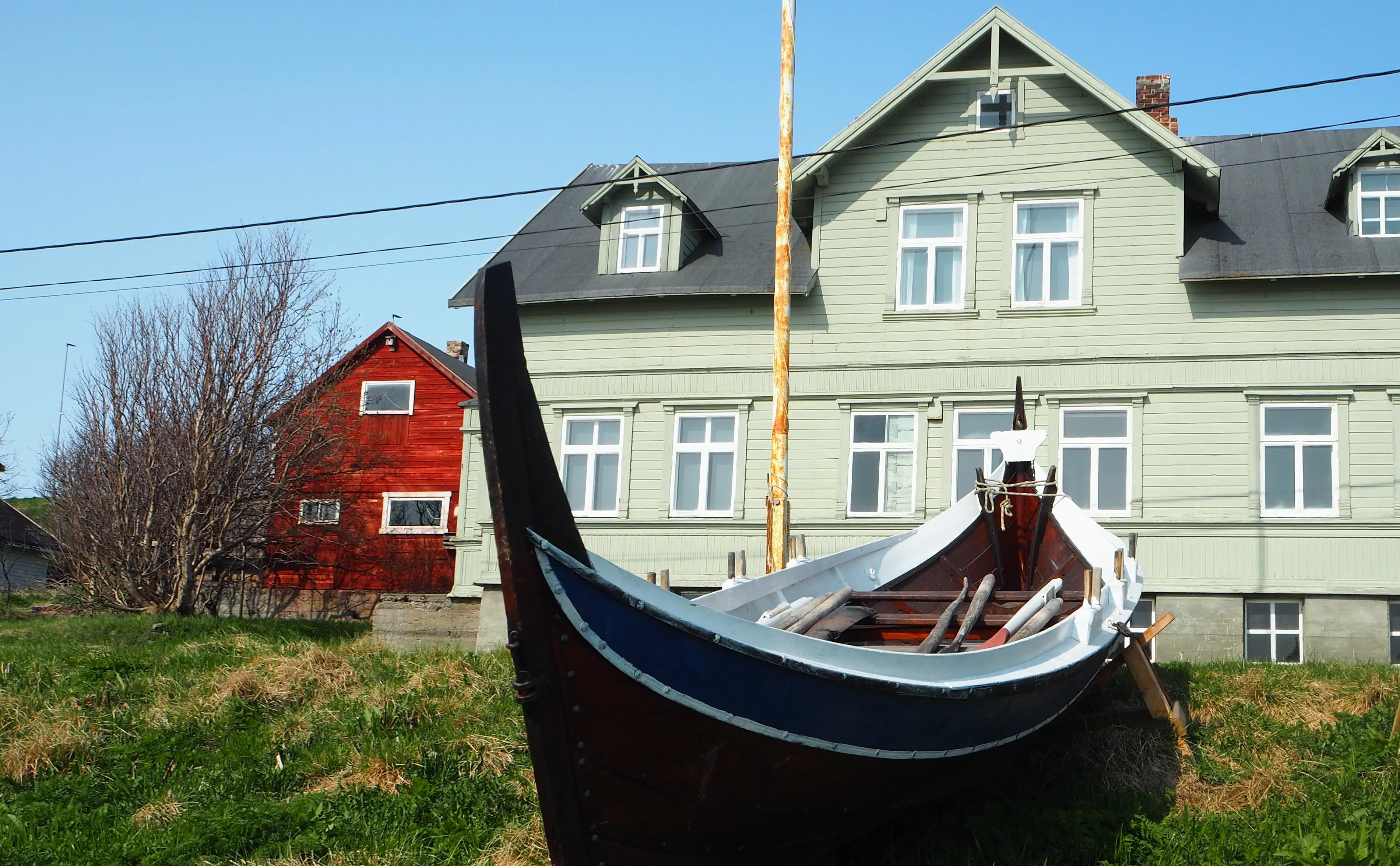 En tradisjonell trebåt med blått og hvitt skrog foran Bietilægårdens grønne hovedbygning og et rødt naboan hus, fotografert på en klar dag