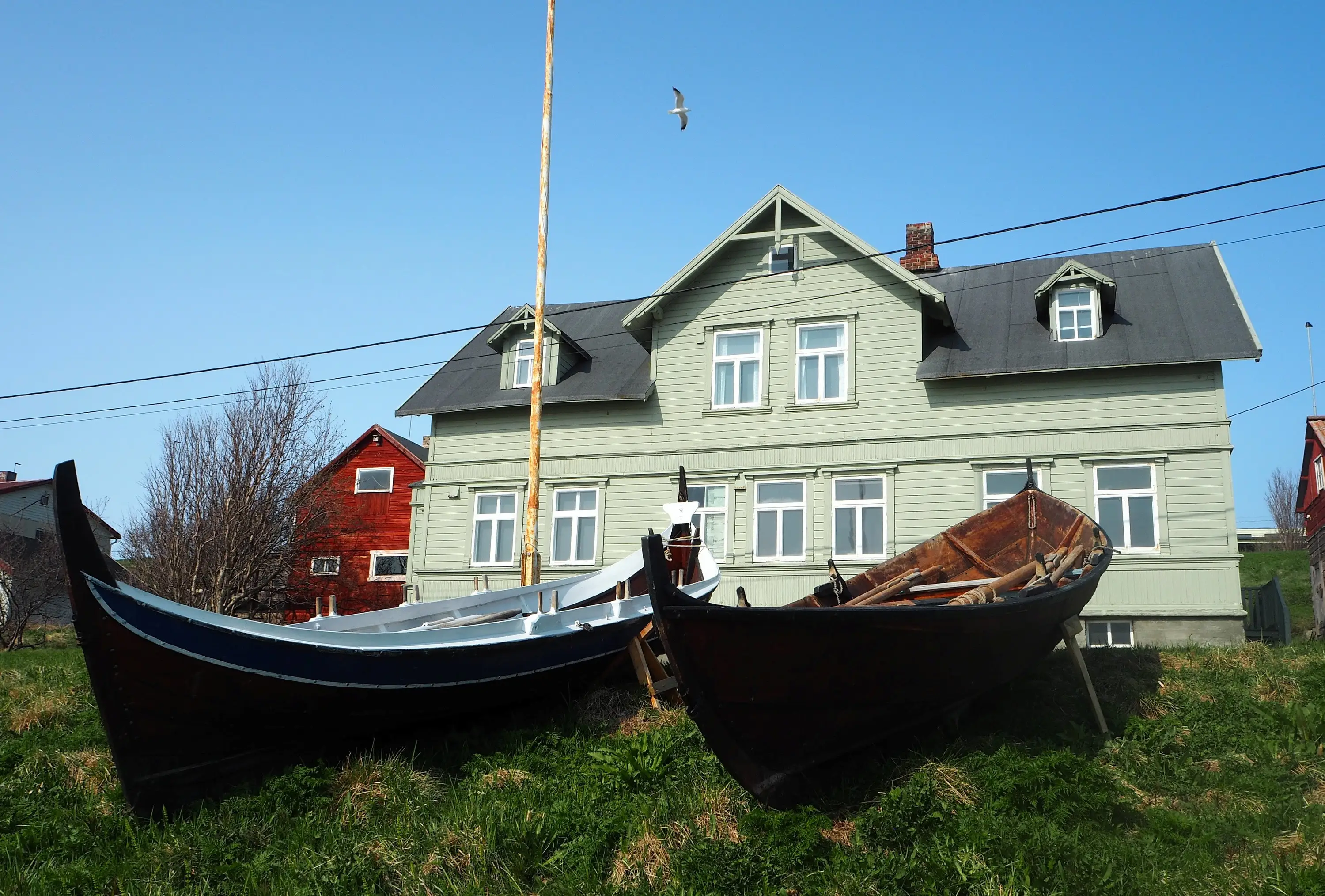 Bietilægården's light green main building with two wooden boats on the lawn in front, on a sunny day