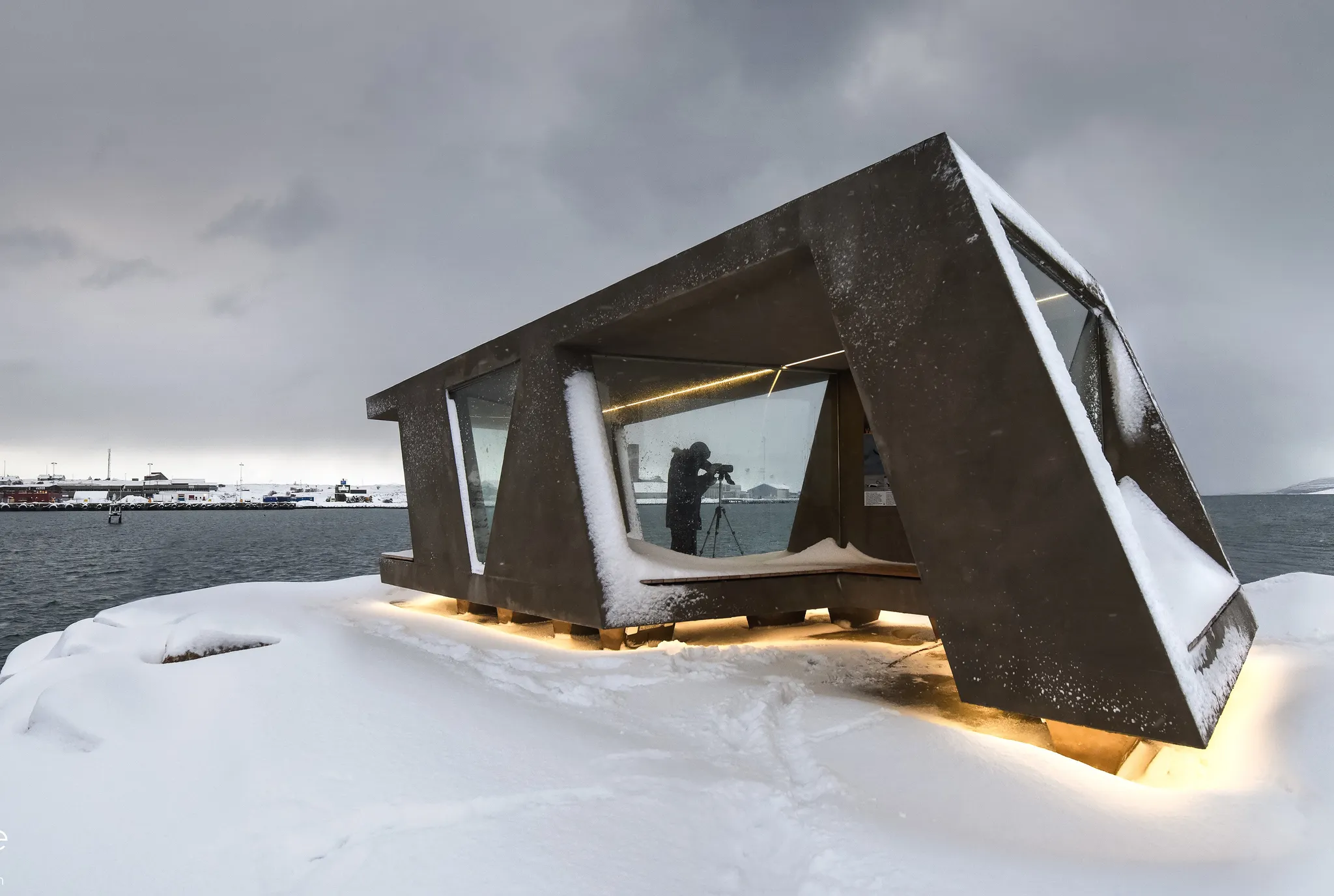 Fuglekikkerskjulet på moloen i Vadsø havn, snødekt en vinterkveld, med en fotograf inne i skjulet