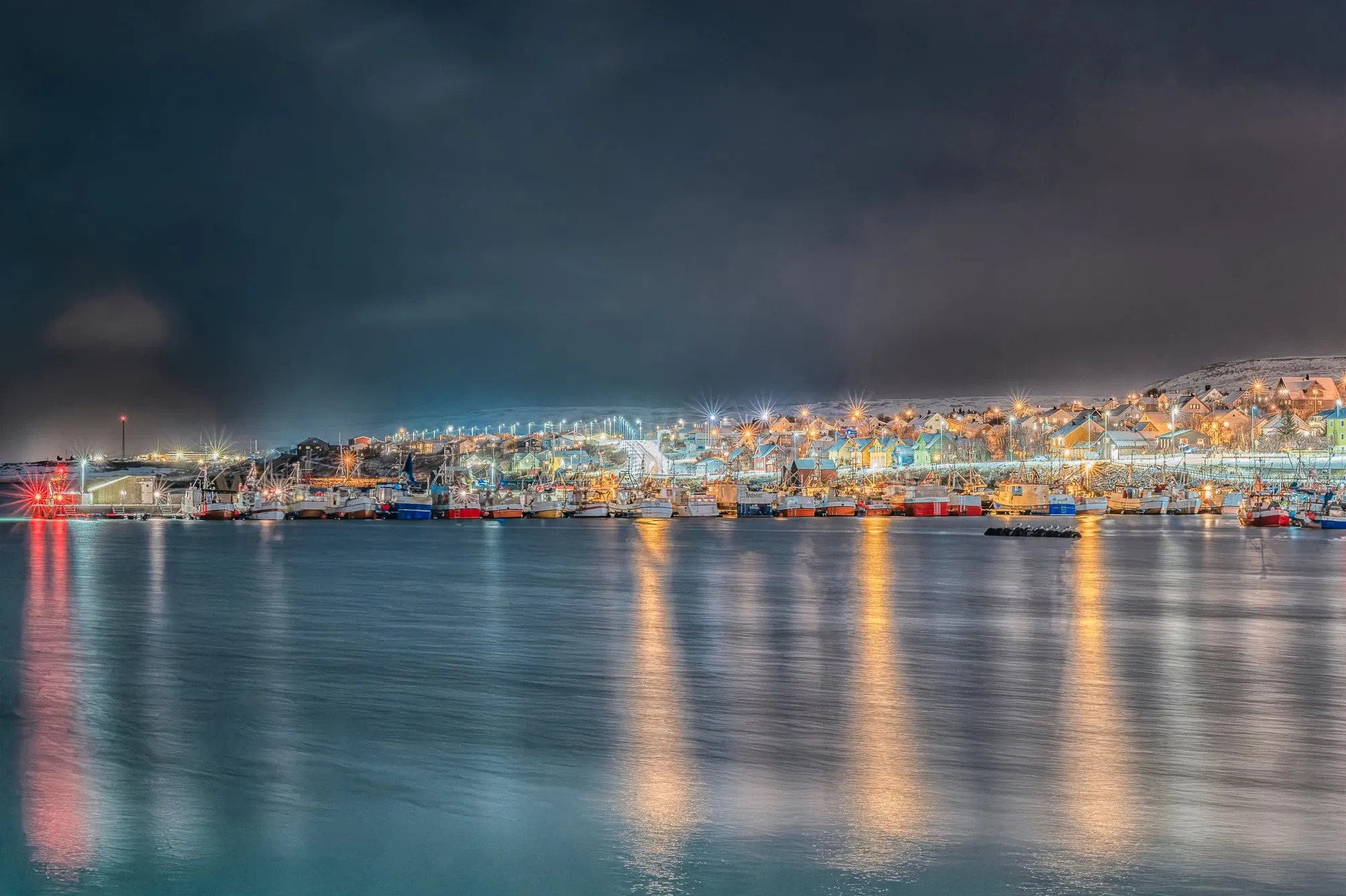 Vadsø town centre and harbour at dusk, with fishing boats along the quay and lights reflected in the water