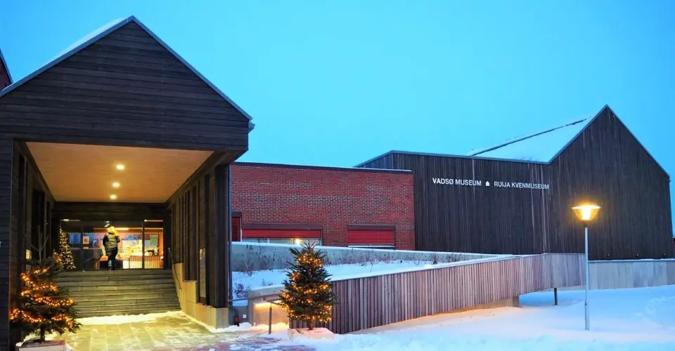 The museum's main building on a winter evening, dark wooden cladding and red brick with the name Vadsø Museum – Ruija Kvenmuseum on the wall, and illuminated Christmas trees by the entrance