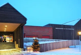 The museum's main building on a winter evening, dark wooden cladding and red brick with the name Vadsø Museum – Ruija Kvenmuseum on the wall, and illuminated Christmas trees by the entrance
