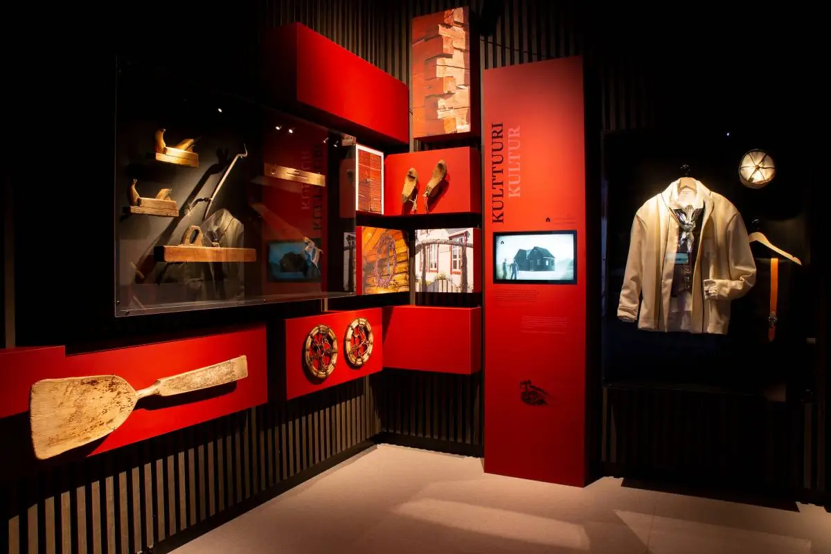 Exhibition room with bright red display walls, Kven work tools and an illuminated white festive costume on a mannequin